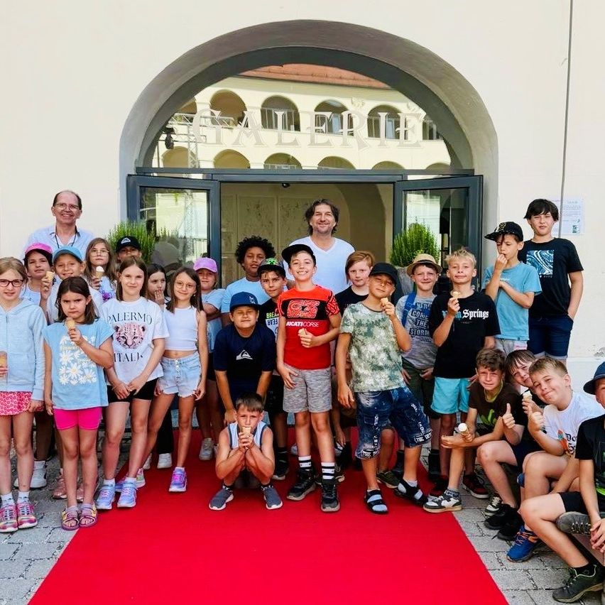 Eine Gruppe von Kindern und Erwachsenen steht auf einem roten Teppich vor einem Gebäude mit einem bogenförmigen Eingang und einem Schild mit der Aufschrift 'PALERE'. Einige Kinder essen Eis und posieren für ein Foto.