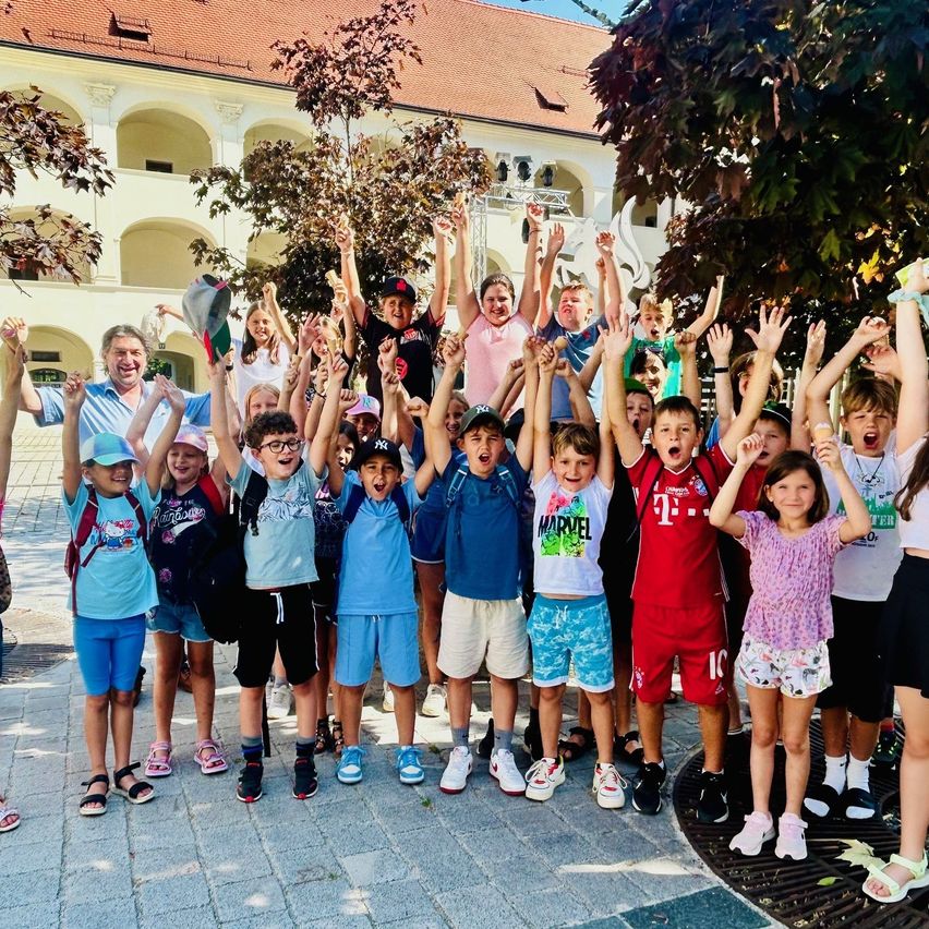 Eine Gruppe von Kindern und Erwachsenen posiert für ein Foto mit erhobenen Händen vor einem Gebäude mit Bogenfenstern und Bäumen.