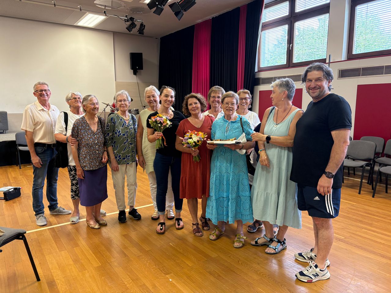 Eine Gruppe älterer Erwachsener steht in einem Raum und lächelt für ein Foto. Einige halten Blumen und einen Teller. Eine Frau in einem blauen Kleid hält einen Kuchen. Der Raum hat Holzböden und eine weiße Wand.