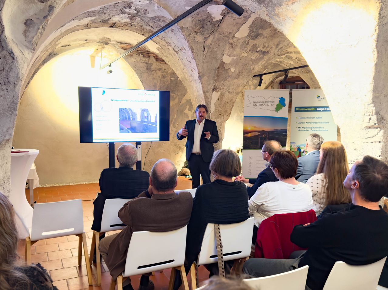 Ein Mann hält eine Präsentation in einem Raum vor einem Publikum, das vor ihm sitzt. Der Präsentationsbildschirm zeigt eine Folie an, und es gibt Banner an den Wänden.