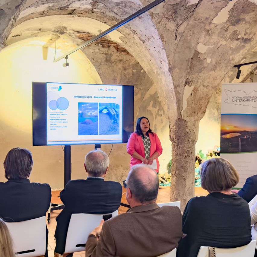 Eine Frau steht vor einer großen Leinwand in einem schwach beleuchteten Raum und präsentiert einen Bericht über Kompost Unterkarnten. Ein Publikum sitzt vor ihr.