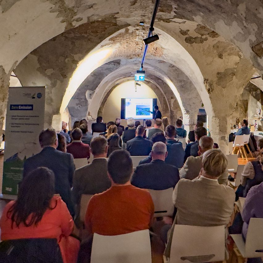 Ein Publikum sitzt in einem gewölbten, bogenförmigen Raum und schaut sich eine Präsentation auf einer Projektionsleinwand an. Ein Schild auf der linken Seite steht 'Null Emission'.