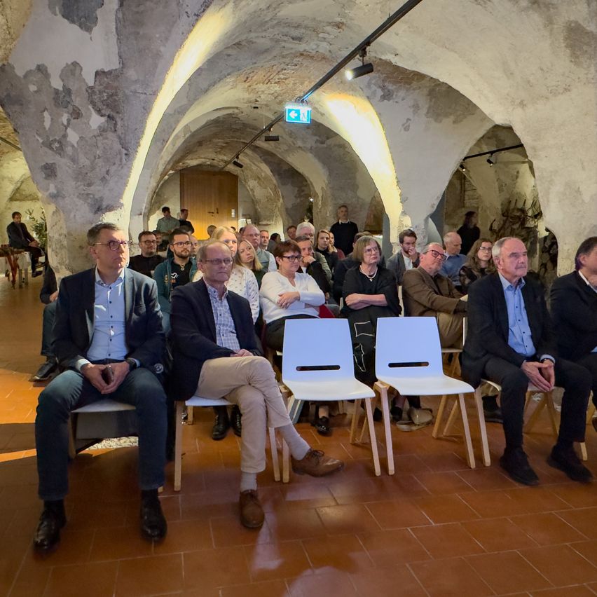 Eine Gruppe von Menschen, meist Männer, sitzt in einem gewölbten Raum. Sie scheinen auf etwas zu warten. Der Raum hat Steinwände und geflieste Böden.