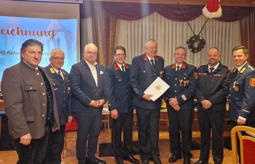 Mehrere Männer in Uniformen und Anzügen stehen zusammen und lächeln für ein Foto. Ein Mann hält ein Dokument. Hinter ihnen zeigt eine Projektionsleinwand ein Bild.