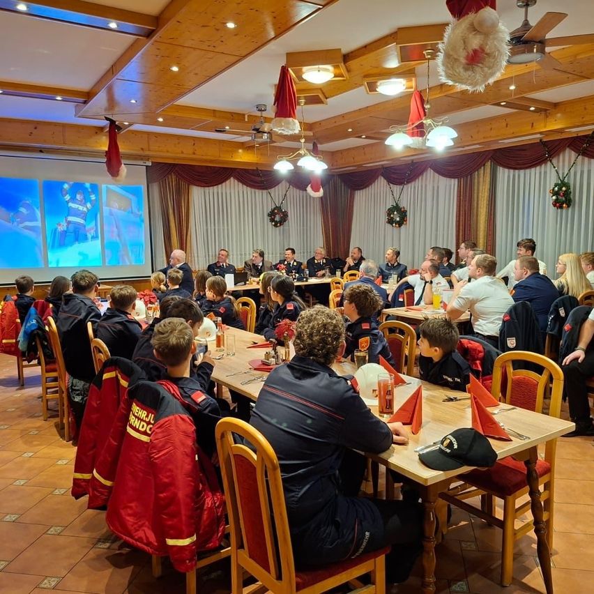 Eine Gruppe von Feuerwehrleuten in Uniformen versammelt sich in einem Raum mit einer Projektionsleinwand, sitzt an langen Tischen. Einige stehen und unterhalten sich. Der Raum ist mit Weihnachtskränzen geschmückt.