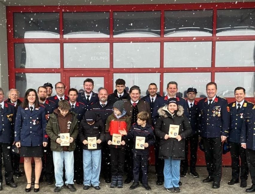 Eine Gruppe von Menschen, darunter Kinder, steht vor einem Gebäude mit roten Fenstern, wahrscheinlich einer Feuerwache. Sie sind alle in Uniformen gekleidet, halten Auszeichnungen in den Händen und lächeln. Das Gebäude ist schneebedeckt.