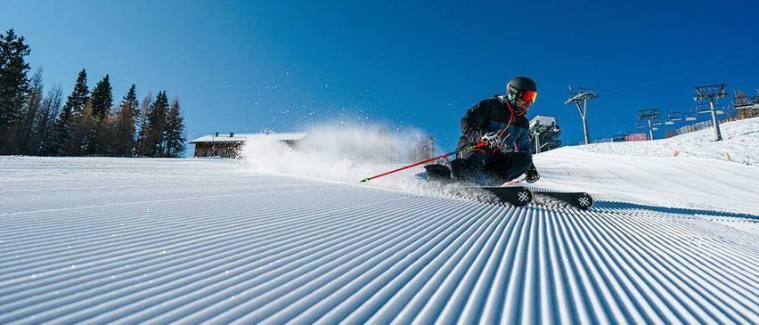 Ein Skifahrer in einer schwarzen Jacke und einem Helm fährt den Schneehang hinunter, mit Bäumen und einem Haus im Hintergrund.