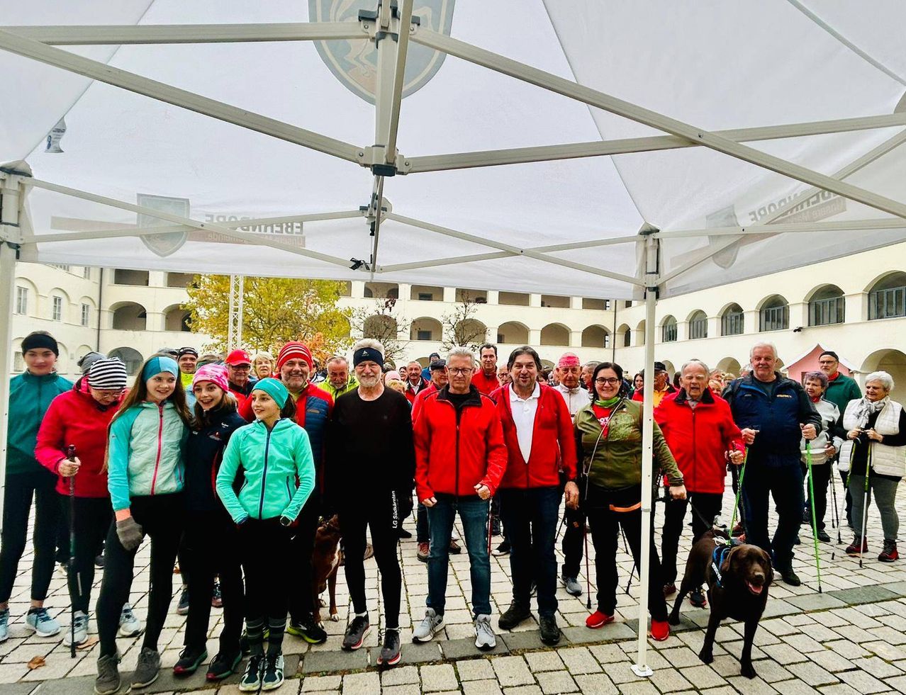Eine Gruppe von Menschen, einschließlich Kindern, steht unter einem Zelt in einem Park. Sie tragen alle rote Jacken. Ein Mann in der Mitte hält einen Hund. Sie alle lächeln.
