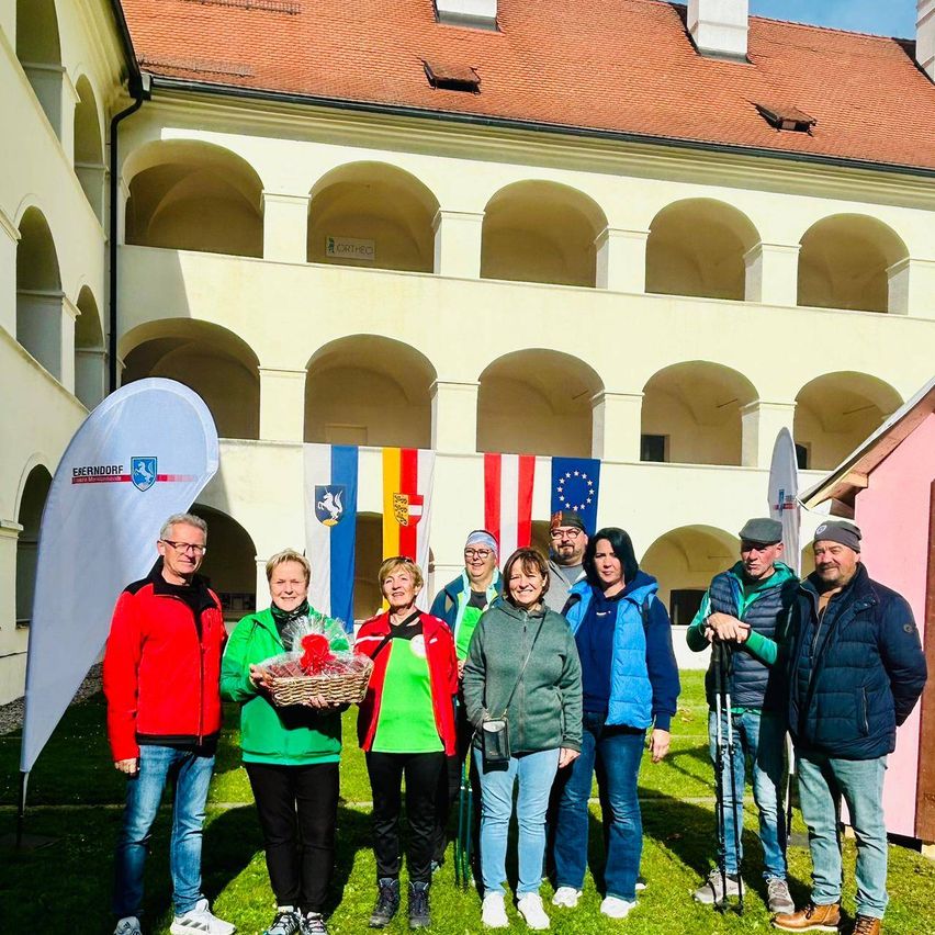Eine Gruppe von Menschen posiert für ein Foto vor einem Gebäude mit Flaggen, eine Person hält einen Korb mit Blumen.
