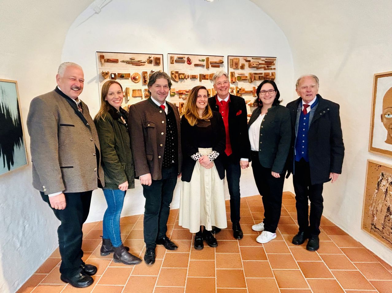 A group of people are standing in front of a wall with various wooden carvings displayed. They are all dressed formally.