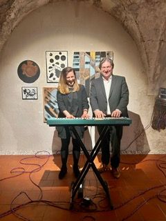 A man and a woman stand before a keyboard, smiling and looking at each other, with various abstract paintings on the wall behind them.