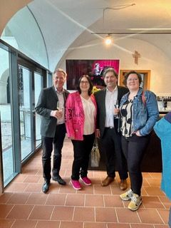 Five people stand in a room with brick floors. They are all smiling for the camera. The woman in the middle wears a pink jacket.