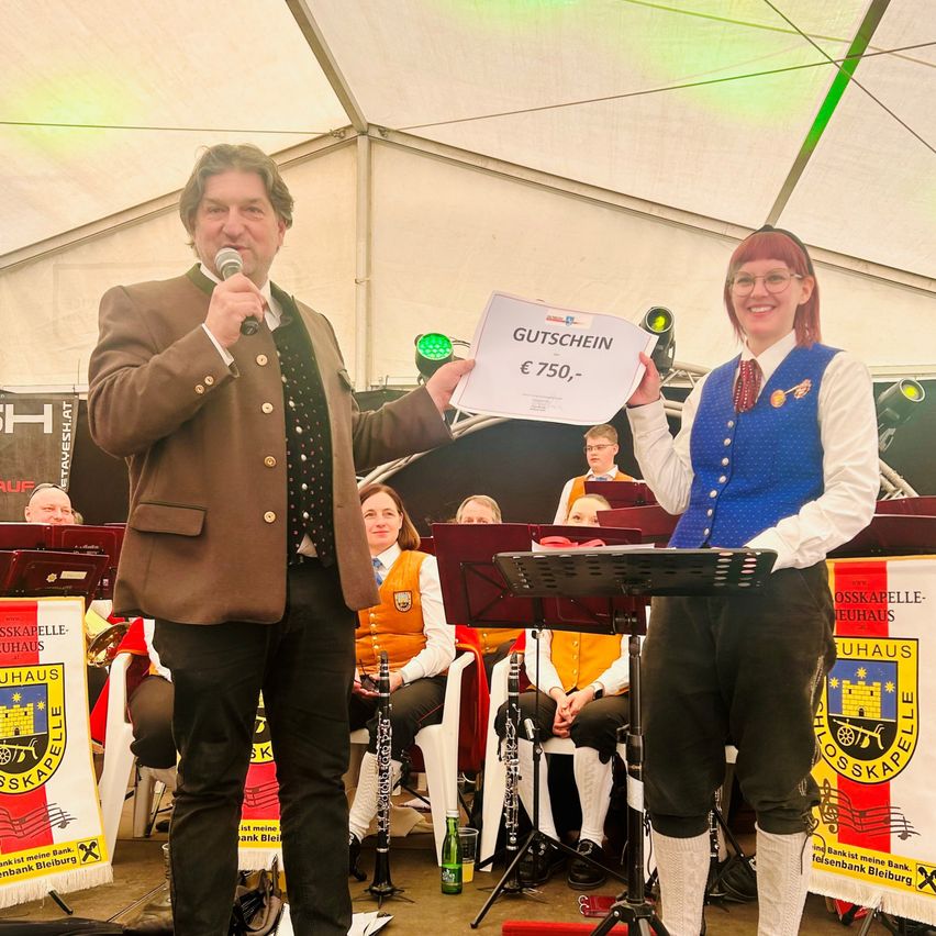 Two people stand on stage, holding a certificate for €750. The man holds a microphone, and the woman smiles. Behind them, a band with instruments sits under a tent. Banners with text are visible in the background.