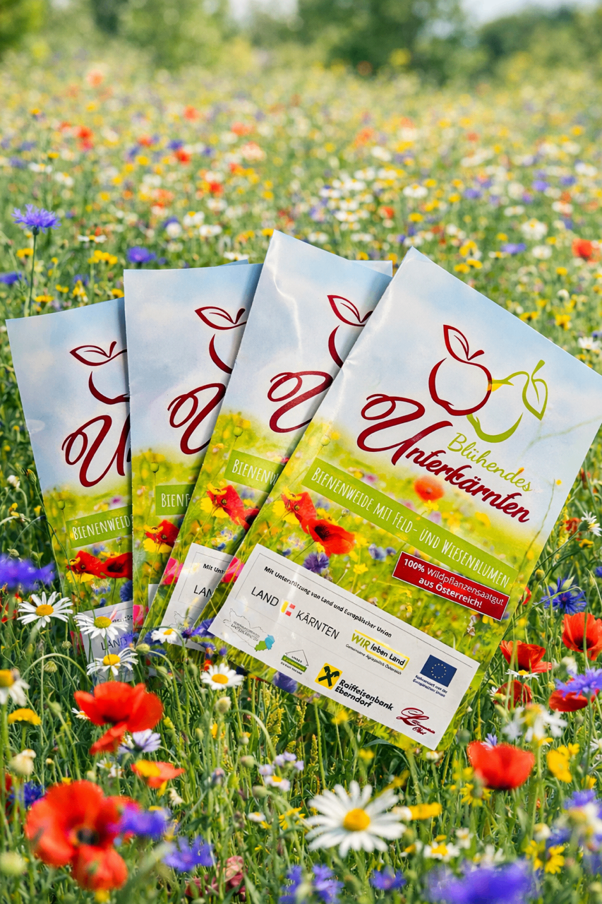 Three packets of seeds labeled 'Bienenweide' are placed on a vibrant field of wildflowers. Each packet has a red apple logo and the words 'Bienenweide mit Feld-und Wiesenblumen aus Österreich!' on it. The packets are positioned in a triangular formation.