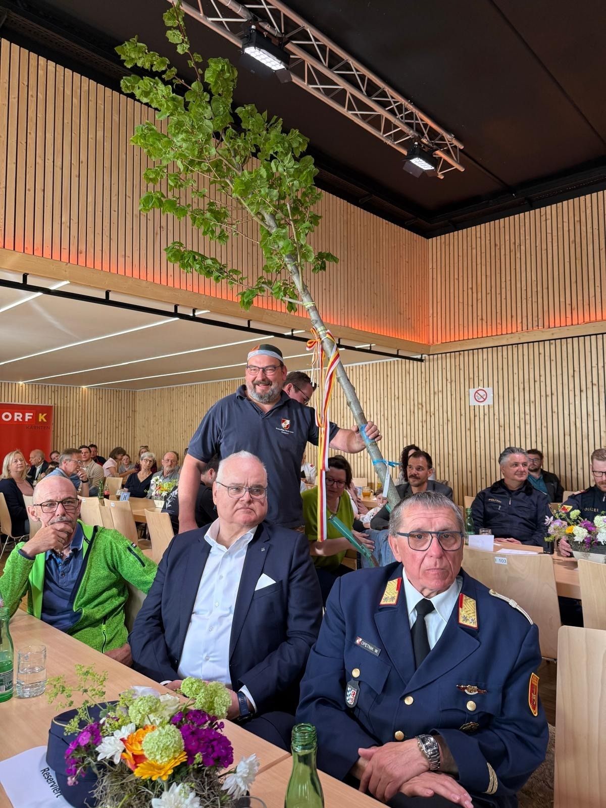 Several men in suits and uniforms sit around a table. A man stands behind them holding a tree with ribbons. Behind them is a wooden wall.