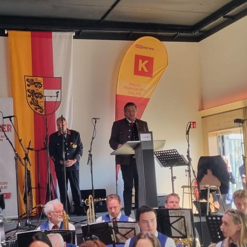 A band is performing in front of an audience. A man stands at a podium with a microphone. Two flags, one with a lion and the other with a yellow K, are displayed behind him.