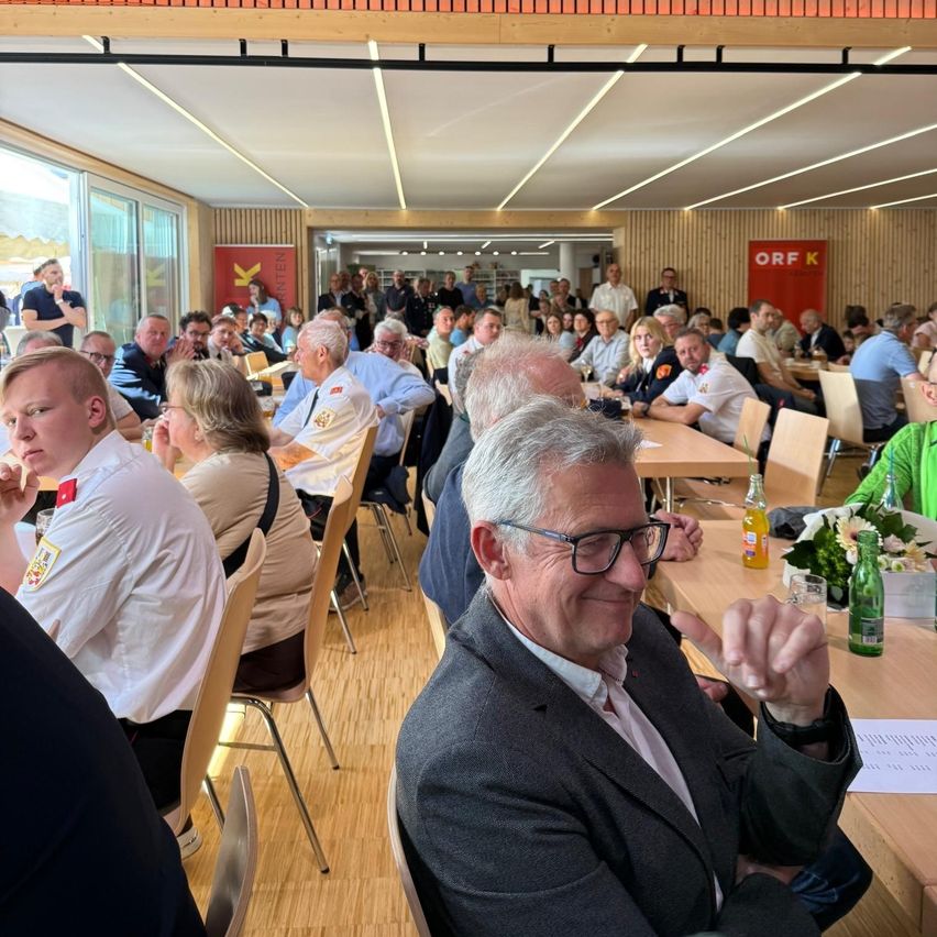 A group of people are seated at tables in a large room, possibly attending an event. A man with gray hair is smiling and making a gesture.
