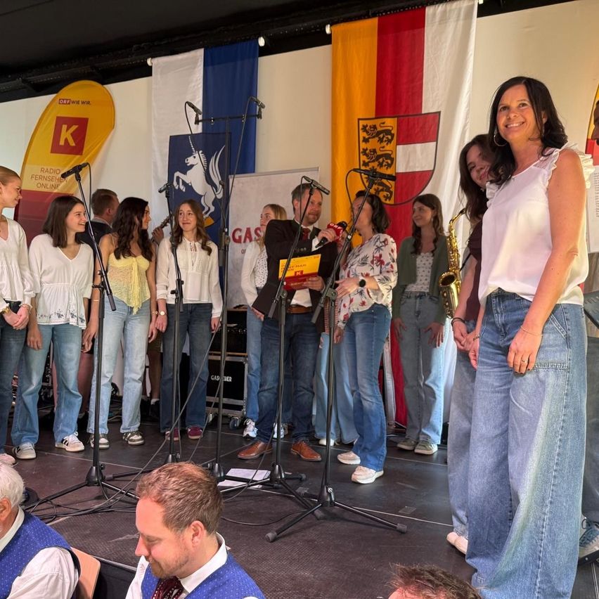 A group of people stand on stage, performing with microphones and a saxophone. Flags hang behind them. A woman in a white blouse smiles.