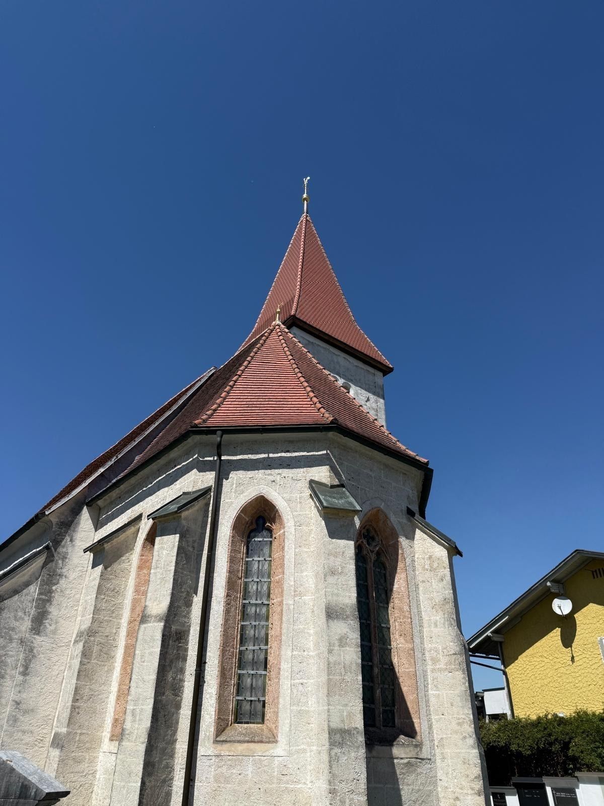 A church with a pointed tower and red roof. Below it, two arched windows are visible. The sky is clear and blue.