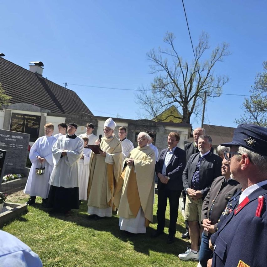 Priests and other men stand on grass with a memorial behind them. They are dressed in formal clothing.