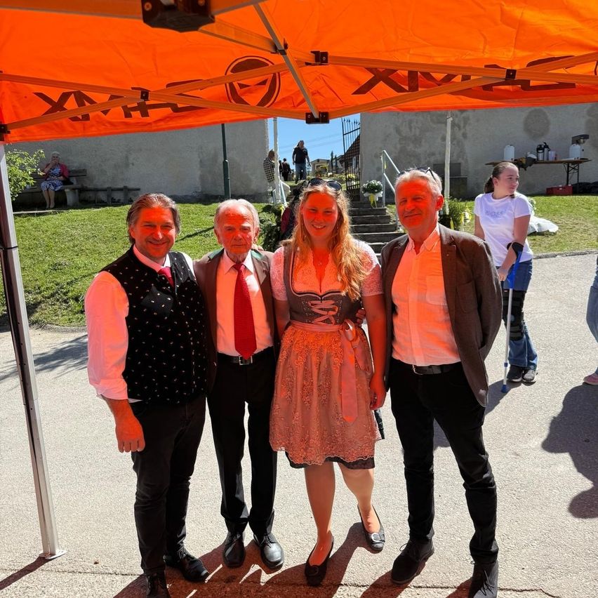 Three men and a woman stand under an orange tent. The woman wears a dirndl, and the men wear suits. They appear to be posing for a photo. Behind them, a girl in a white shirt is walking.