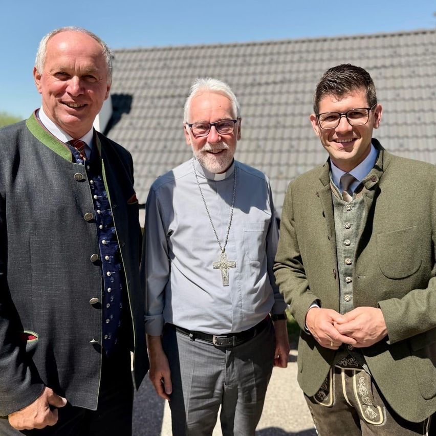 Three men stand together outside. The man on the left wears a traditional green jacket. The middle man wears a necklace with a cross. The man on the right wears glasses.