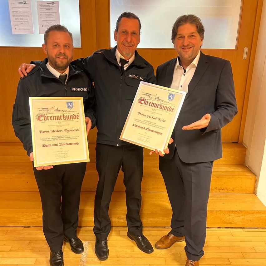 Three men stand together, smiling and holding certificates. The man on the left wears a police uniform, while the other two are in suits. The man in the middle holds a certificate with the words 'Chemarkunde' on it. The room has wooden floors and a glass wall.