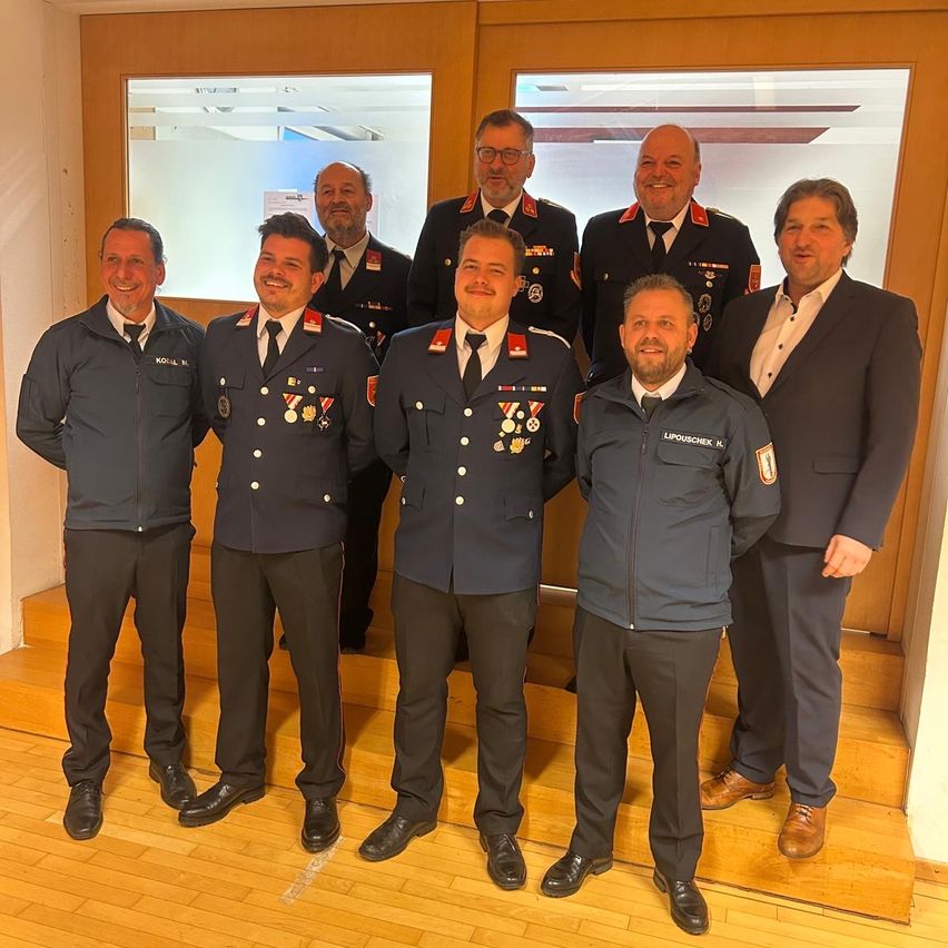 Seven men in uniforms stand together in a room, smiling. They wear medals and appear to be posing for a photo. The room has wooden floors and brown walls.