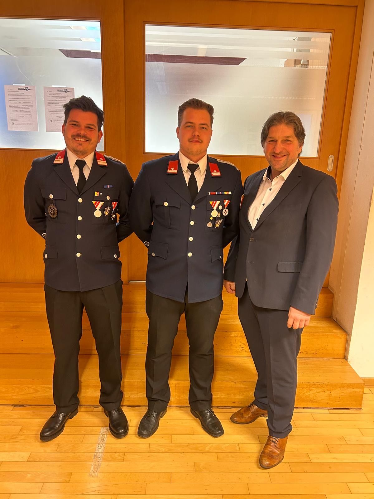 Three men stand in front of a door, all wearing suits. Two of them are in uniform, adorned with medals. They are smiling and seem to be posing for a photo.
