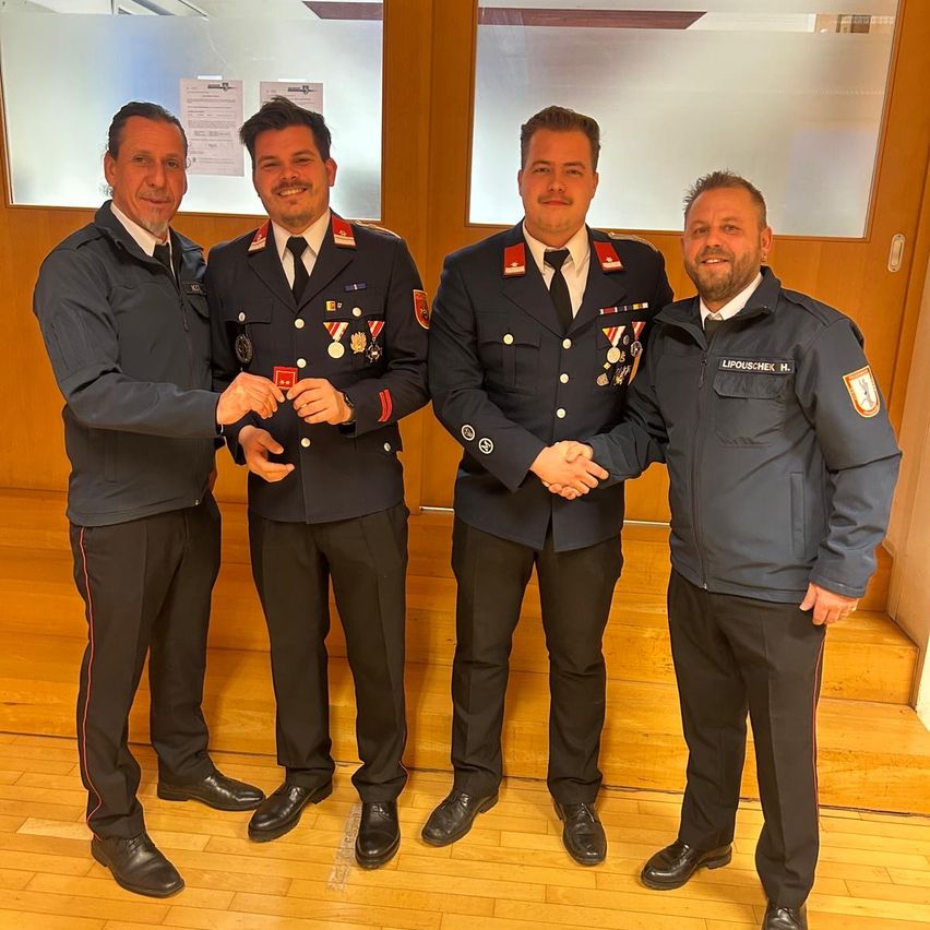 Four men in uniforms are standing in a room, smiling. The man on the left is giving a medal to the man in the middle. The man on the right is holding the hand of the man in the middle. Behind them, there is a wooden door with a glass panel.