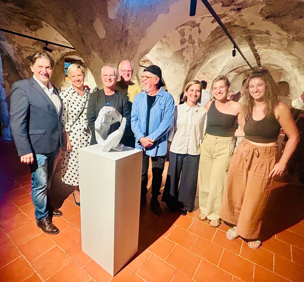 Eine Gruppe von Menschen steht um einen weißen Sockel mit einer Glasplastik darauf. Sie alle lächeln und scheinen für ein Foto zu posieren.