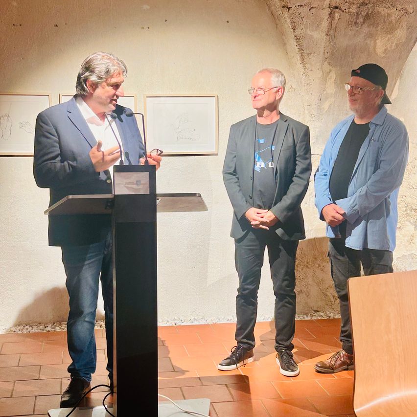 Drei Männer stehen in einem Raum mit gefliesten Böden und einer Steinwand. Ein Mann spricht in ein Mikrofon an einem Podium, während die anderen beiden zuhören.