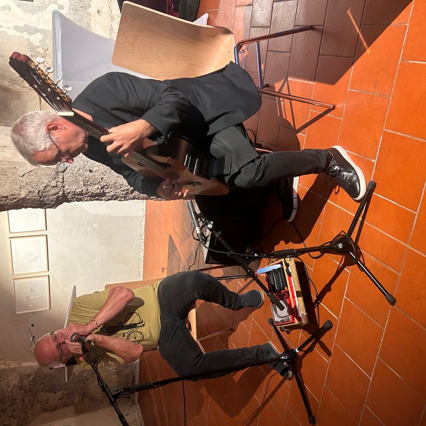 Zwei Männer spielen auf einem gefliesten Boden, einer spielt Gitarre und der andere singt in ein Mikrofon.