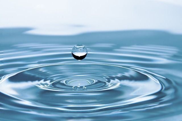 Ein Wassertropfen schwebt in der Luft und erzeugt Wellen auf der Wasseroberfläche. Der Tropfen reflektiert den Himmel.