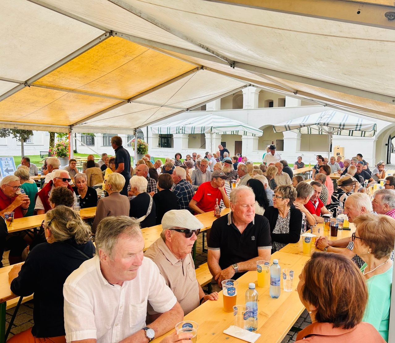 Eine große Gruppe von Menschen sitzt unter einem Zelt an Tischen, möglicherweise bei einem Festival. Viele tragen Hüte und Sonnenbrillen, und einige halten Gläser mit Bier. Die Tische sind mit verschiedenen Gegenständen gefüllt, darunter Flaschen und Tassen. Im Hintergrund befindet sich ein Gebäude mit Säulen.