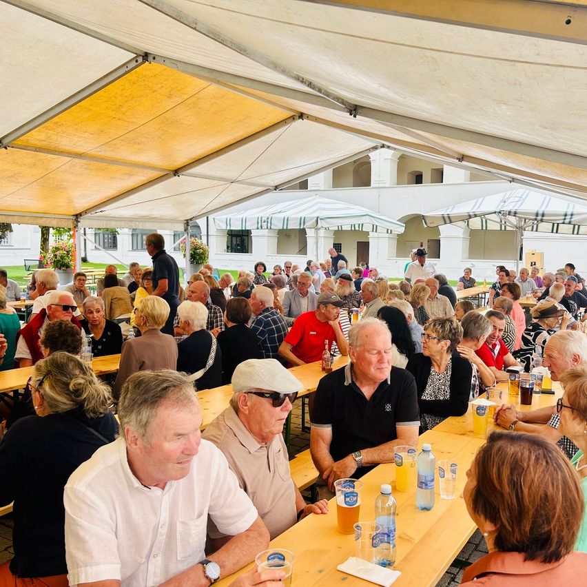 Eine große Gruppe von Menschen sitzt unter einem Zelt an Tischen und genießt Getränke und Essen. Die Tische sind mit Gläsern, Flaschen und Servietten gefüllt. Die Umgebung scheint ein Festival oder eine Veranstaltung im Freien zu sein.