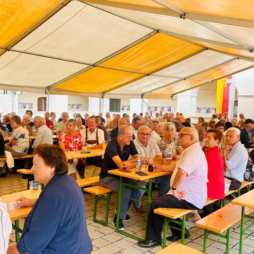 Eine große Gruppe von Menschen, überwiegend ältere Personen, sitzen unter einem gelben Zelt an Tischen. Einige unterhalten sich, während andere umherschauen. Die Tische sind mit Flaschen, Gläsern und anderen Gegenständen bestückt. Es scheint sich um ein gesellschaftliches Treffen oder eine Veranstaltung zu handeln.