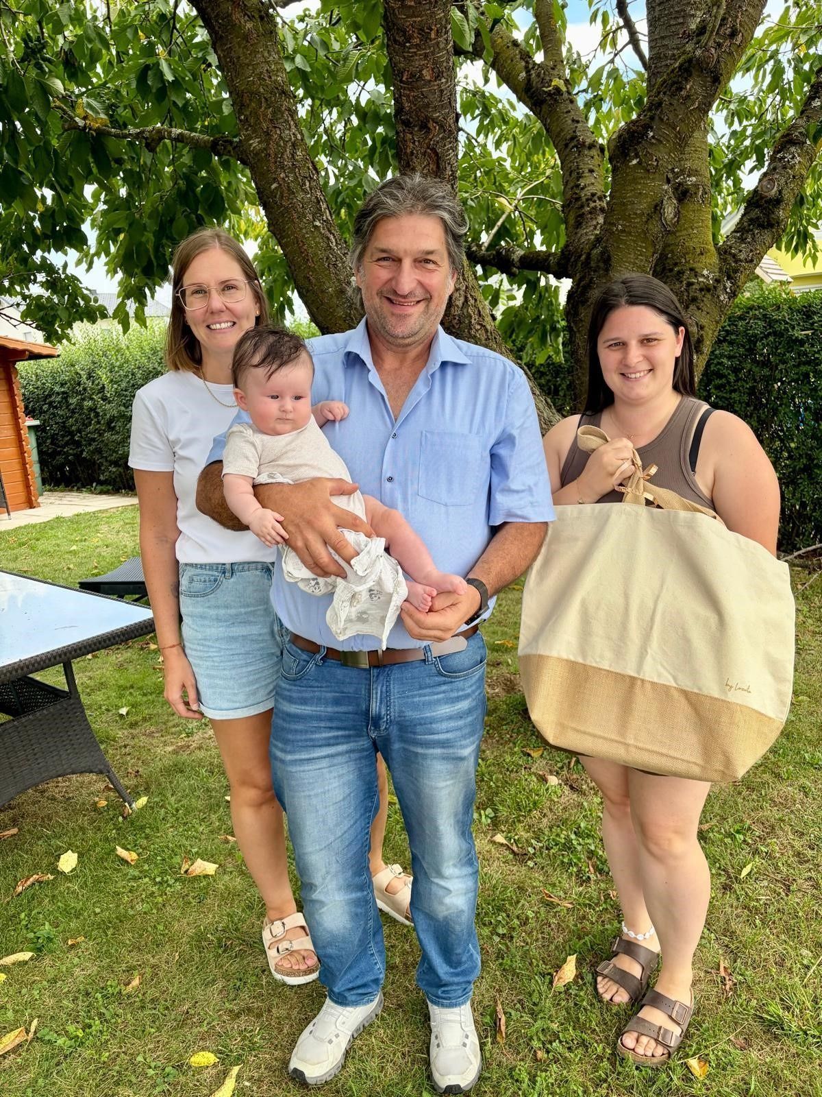 Eine dreiköpfige Familie, bestehend aus einem Mann, der ein Baby hält, einer Frau, die eine Tragetasche hält, und einer weiteren Frau, die hinter ihnen steht, alle lächelnd und posierend für ein Foto im Garten.