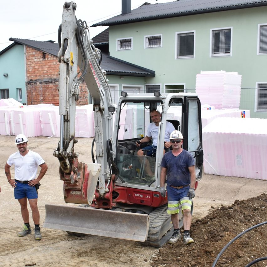 Drei Männer sind auf einer Baustelle mit einem roten Bagger. Ein Mann sitzt im Bagger, während die anderen beiden auf der linken Seite stehen. Dahinter sind gestapelte rosa Objekte und im Hintergrund sind Gebäude.