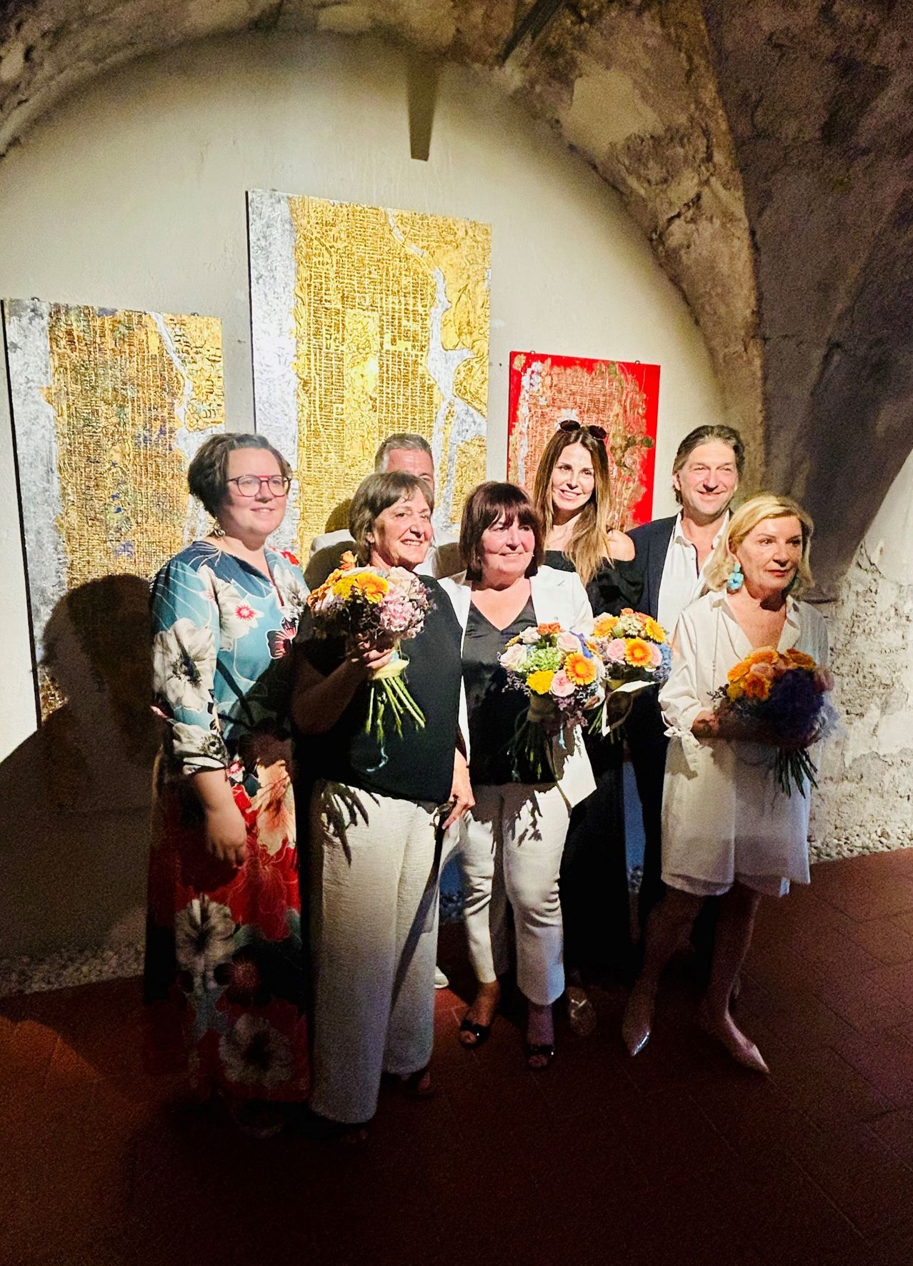 Sieben Personen stehen eng beieinander in einem Raum mit Kunst an den Wänden. Sie halten Blumensträuße und schauen in die Kamera. Die Frau in der Mitte trägt eine Brille.