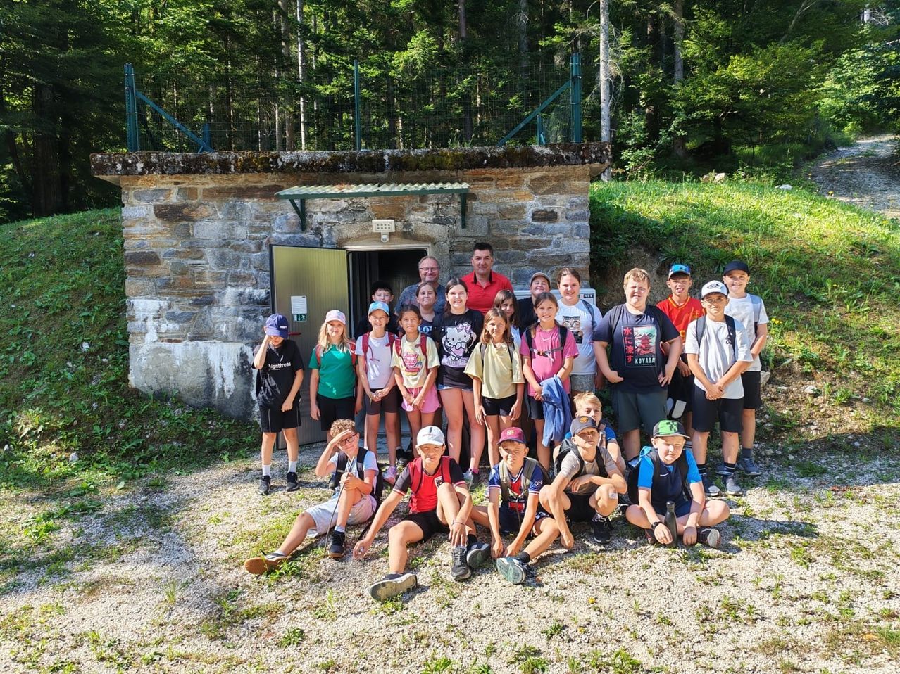 A group of children and adults pose for a photo outside a stone building with an open door. They are on a gravel ground with a grassy area in the background.
