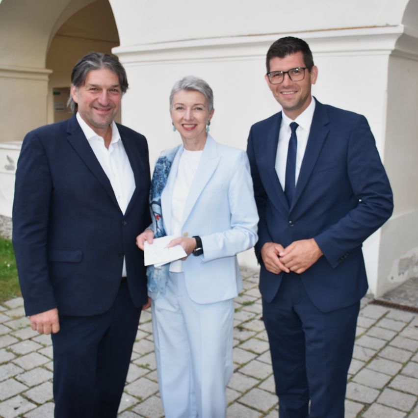 Three formally dressed individuals stand in front of a building. The woman in the center holds a piece of paper. The man on the left smiles, while the man on the right looks at the camera.