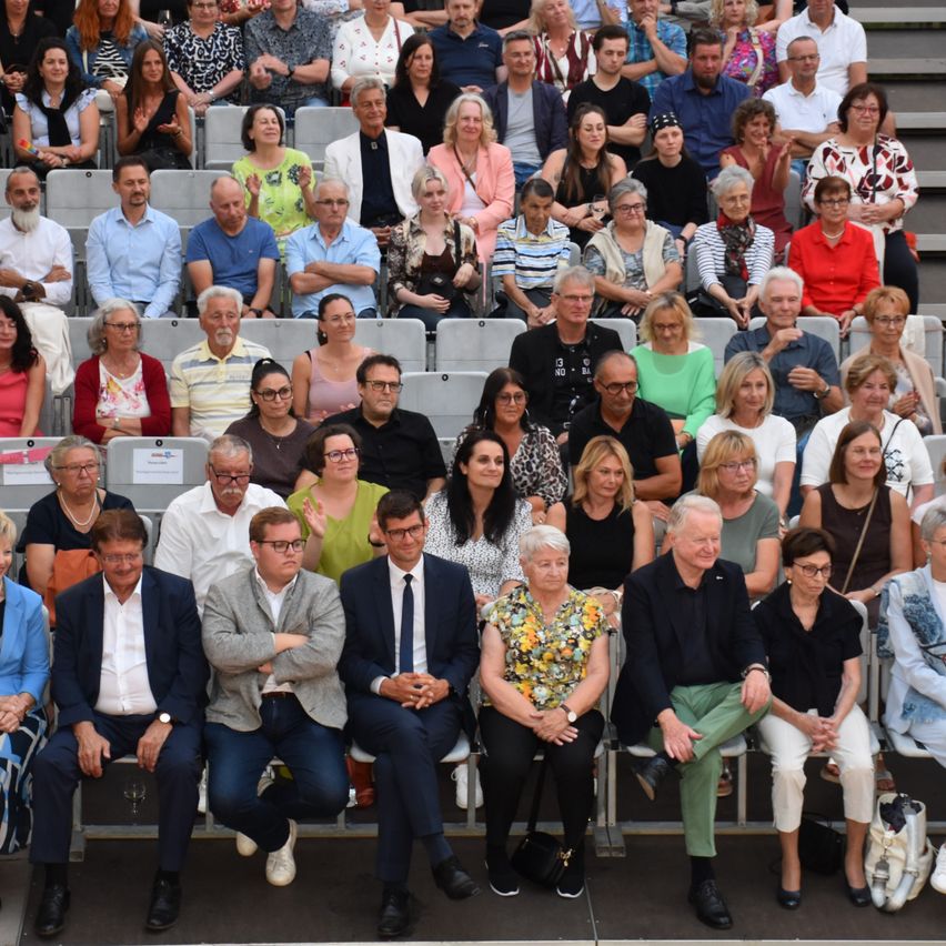 A diverse group of people, possibly attendees of an event, are seated in rows of chairs. Some are wearing glasses, and a few have handbags. They are looking in various directions.