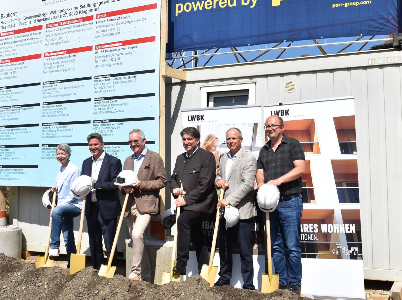 A group of men and women stand in front of a construction site, holding shovels and wearing hard hats. A large banner behind them reads 'powered by'.