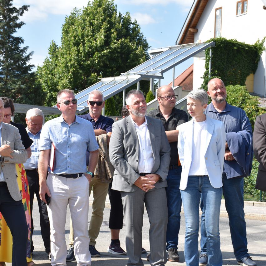 A group of people stands in front of a house with solar panels, some wearing sunglasses and casual attire.