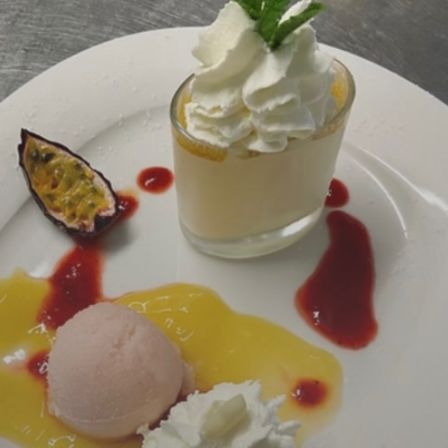 Ein weißer Teller mit einer Vanillecreme, die mit Schlagsahne und einer Minzblatt gekrönt ist, begleitet von einer Kugel Eiscreme, einer Passionsfruchtscheibe und einer Himbeersauce.
