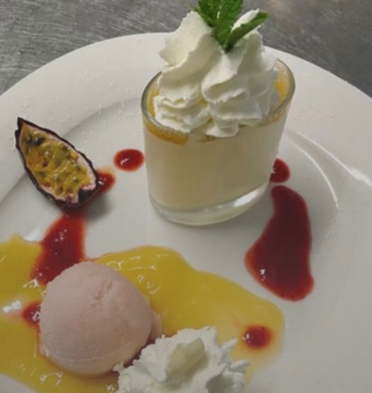 A white plate with a vanilla custard topped with whipped cream and a mint leaf, accompanied by a scoop of ice cream, a passion fruit slice, and a raspberry sauce.