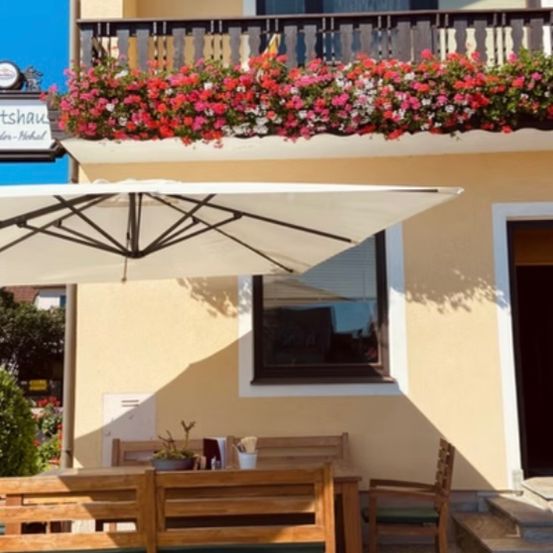 An outdoor dining area with a large umbrella and a table with chairs in front of a building. The building has a balcony with a railing and flowers.