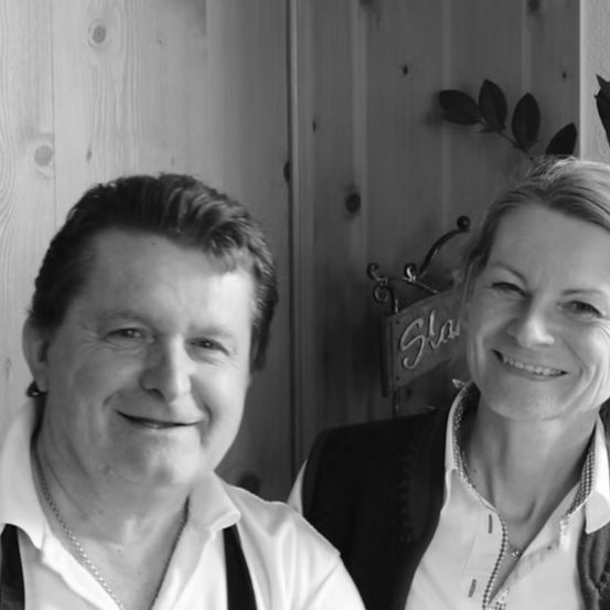 A smiling man and woman stand close together, with the man wearing a necklace and suspenders and the woman wearing a blazer. They are positioned against a wooden wall with a decorative sign behind them.