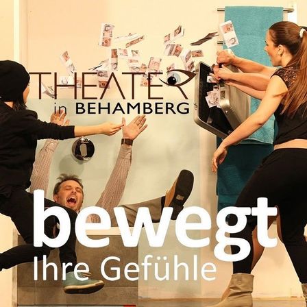 Eine Gruppe von Menschen führt ein Theaterstück in Behamberg auf. Ein Mann liegt auf dem Boden, während zwei Frauen Geld und eine offene Tasche handhaben. An der Wand hinter ihnen befindet sich ein großes Auge und die Worte 'Theater in Behamberg'.